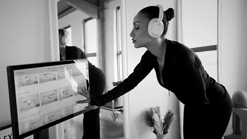 Studio de Pilates Reformer digital en noir et blanc : une cliente sélectionne sa séance sur un kiosque tactile près des Reformers