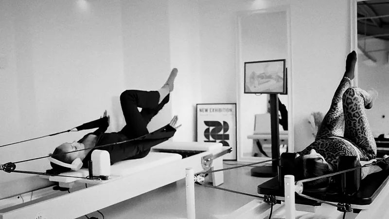 Studio de Pilates Reformer digital en noir et blanc : deux pratiquantes sur Reformer, kiosque tactile affichant la plateforme Reformer Society Connect
